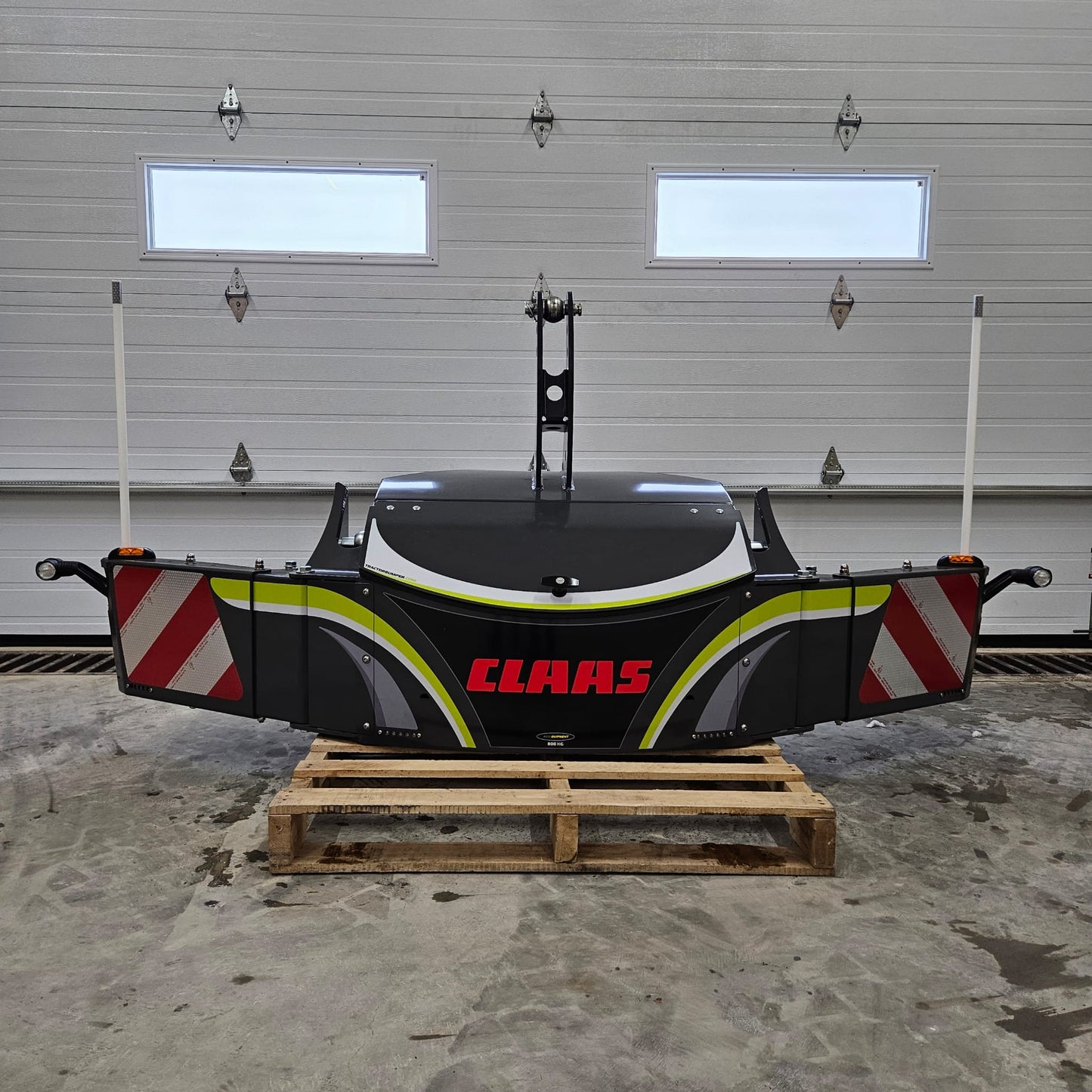 A large black and gray CLAAS TractorBumper 800KG rests on a wooden pallet in a garage with a closed white overhead door and two small windows, featuring integrated LED lighting for enhanced visibility.