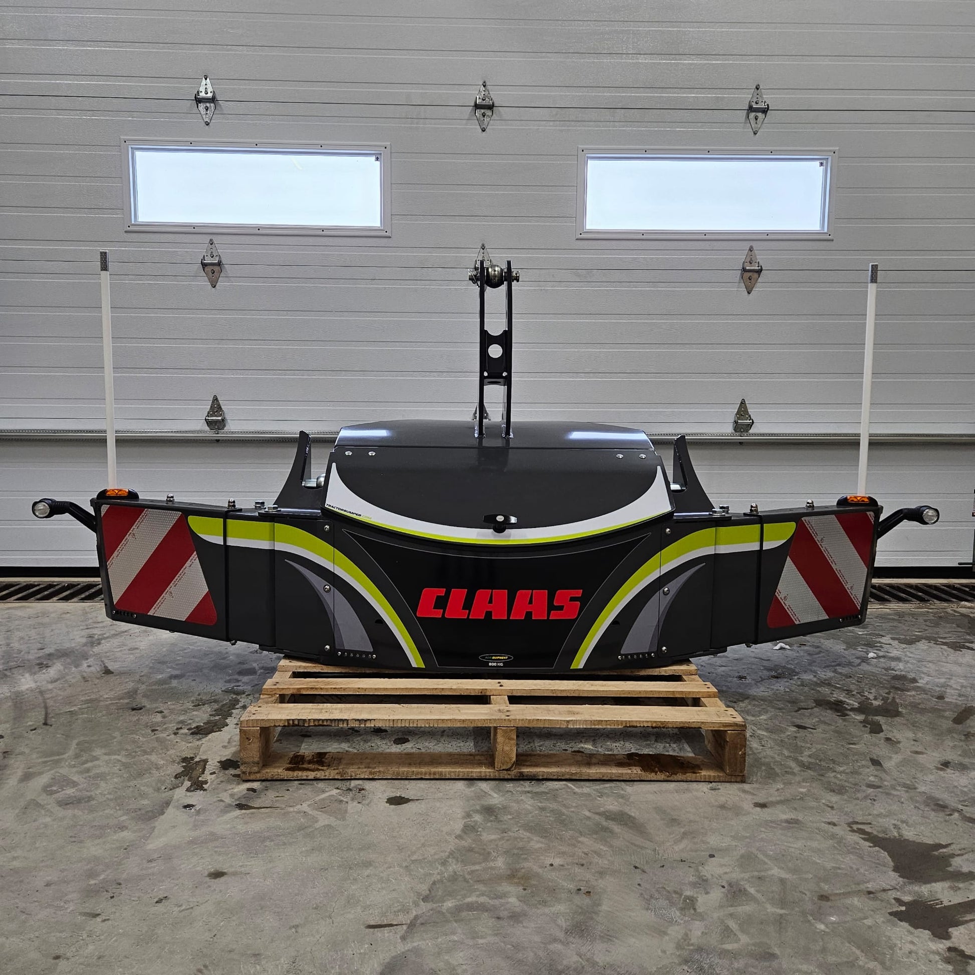 A large black and gray CLAAS TractorBumper 800KG rests on a wooden pallet in a garage with a closed white overhead door and two small windows, featuring integrated LED lighting for enhanced visibility.