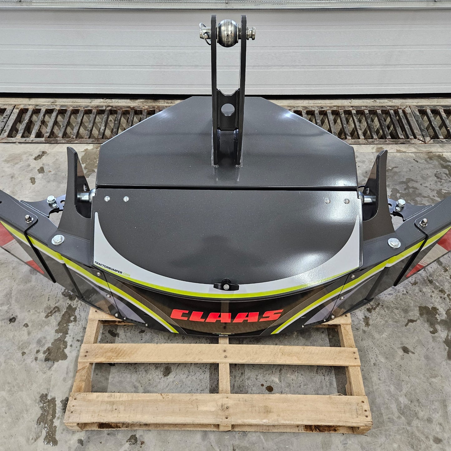 A CLAAS TractorBumper 800KG, gray in color, is placed on a wooden pallet indoors on a concrete floor. The clean agricultural attachment is viewed from above, with a large metal door visible in the background.
