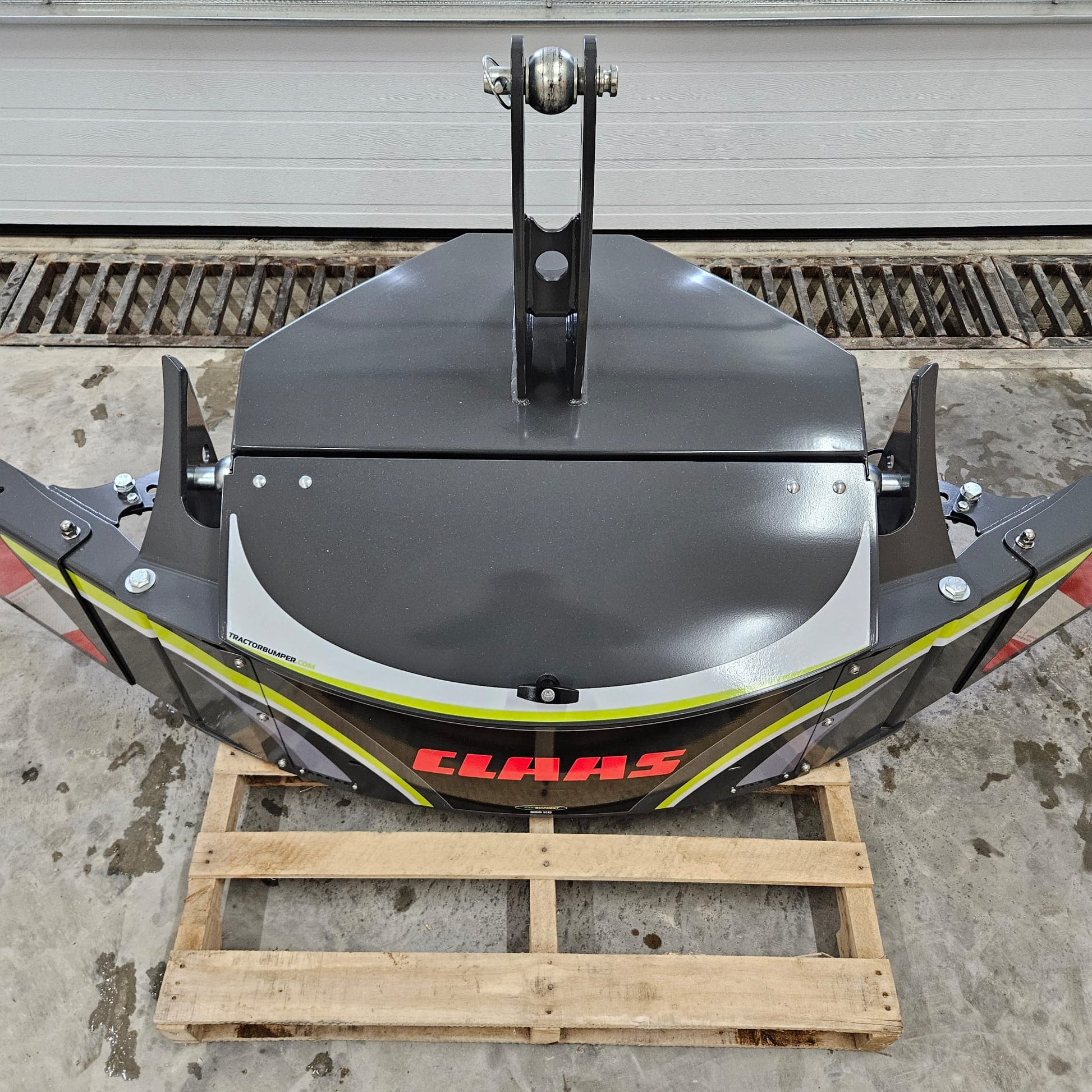 A CLAAS TractorBumper 800KG, gray in color, is placed on a wooden pallet indoors on a concrete floor. The clean agricultural attachment is viewed from above, with a large metal door visible in the background.