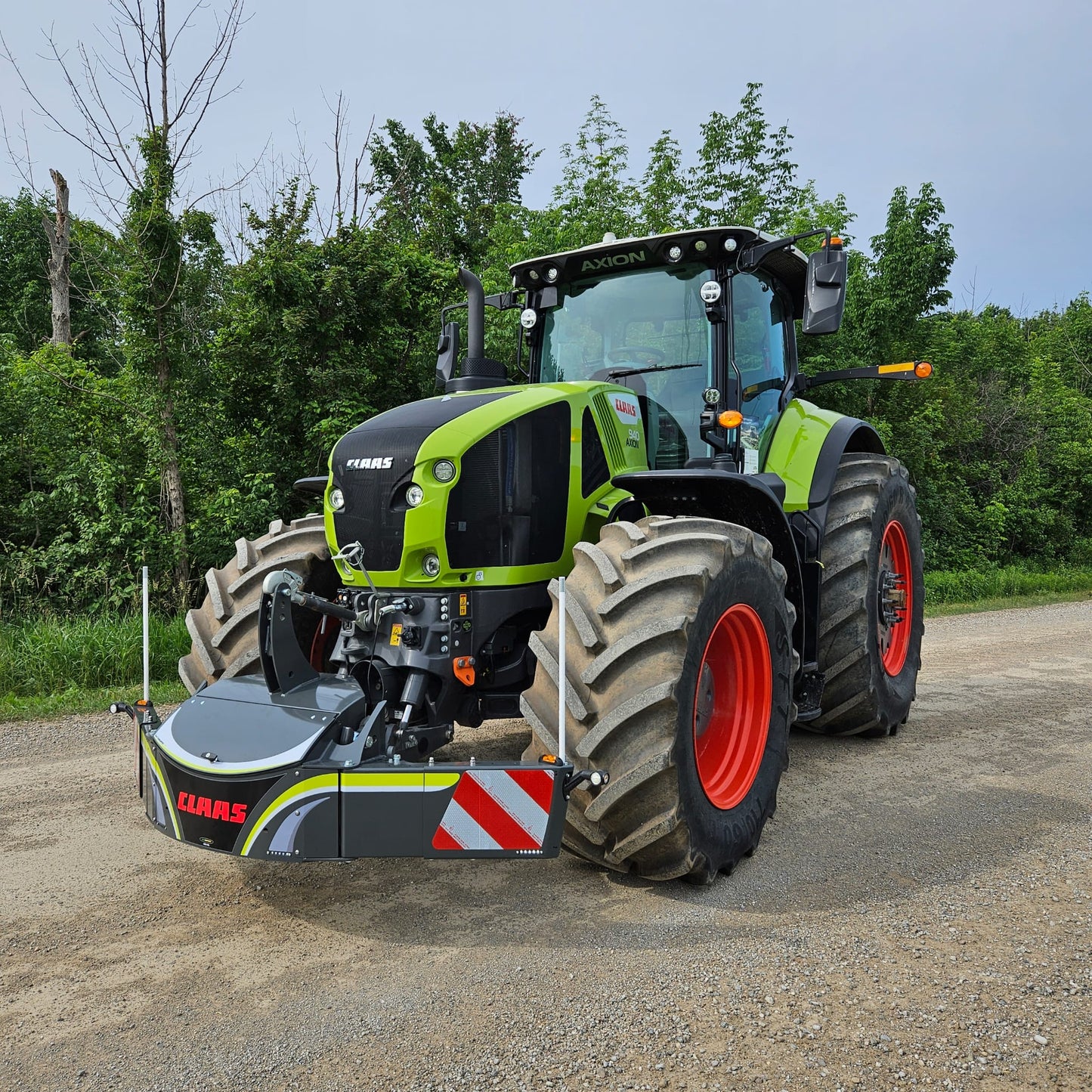 A CLAAS tractor equipped with a TractorBumper 800KG accessory is parked on a dirt road, surrounded by lush green trees and vegetation in the background.