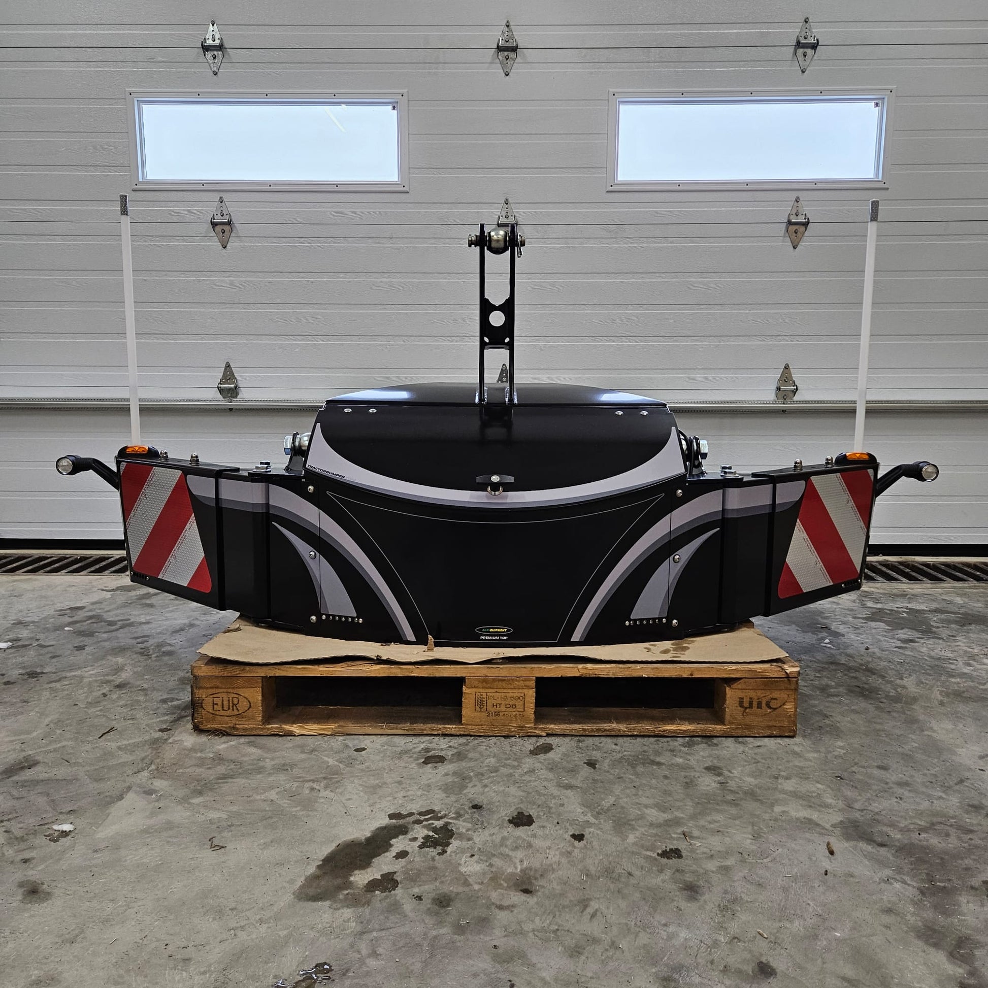 The TractorBumper Toolbox, a spacious black XL sealed toolbox with red-and-white striped panels, is placed on a wooden pallet in a garage with a white sectional door. Two white poles and integrated LED lighting enhance its functionality.