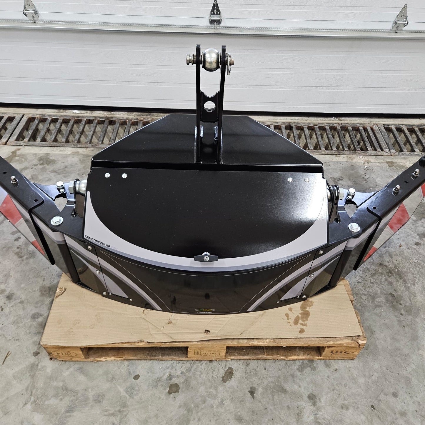 A black and silver industrial metal TractorBumper Toolbox is mounted on a wooden pallet indoors, with a white garage door and concrete floor in the background.
