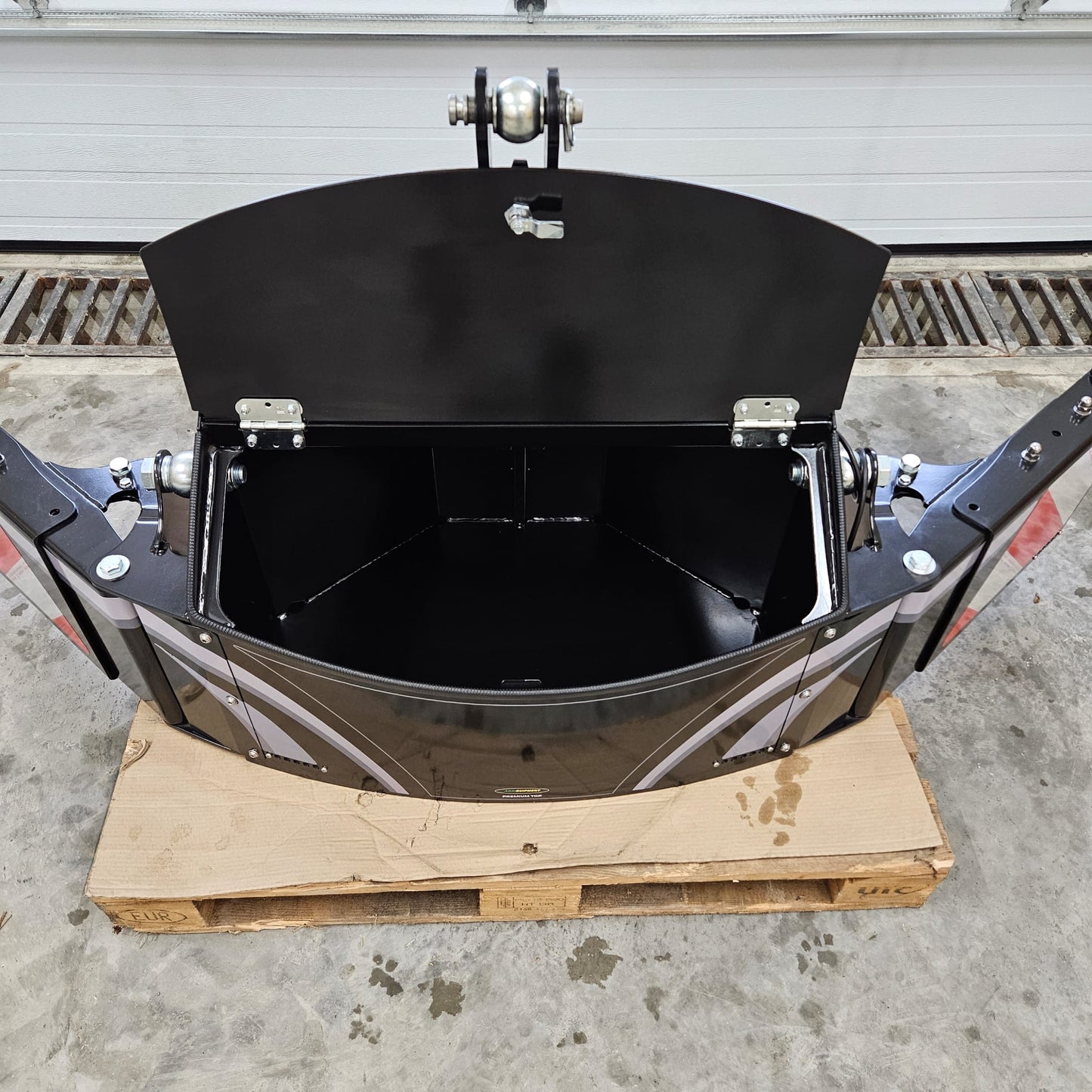 A black XL TractorBumper Toolbox with a hinged lid open, mounted on a wooden pallet in a garage. Features metal hinges and bolts, red side accents, and sits on a concrete floor—ideal for secure storage in your workspace.