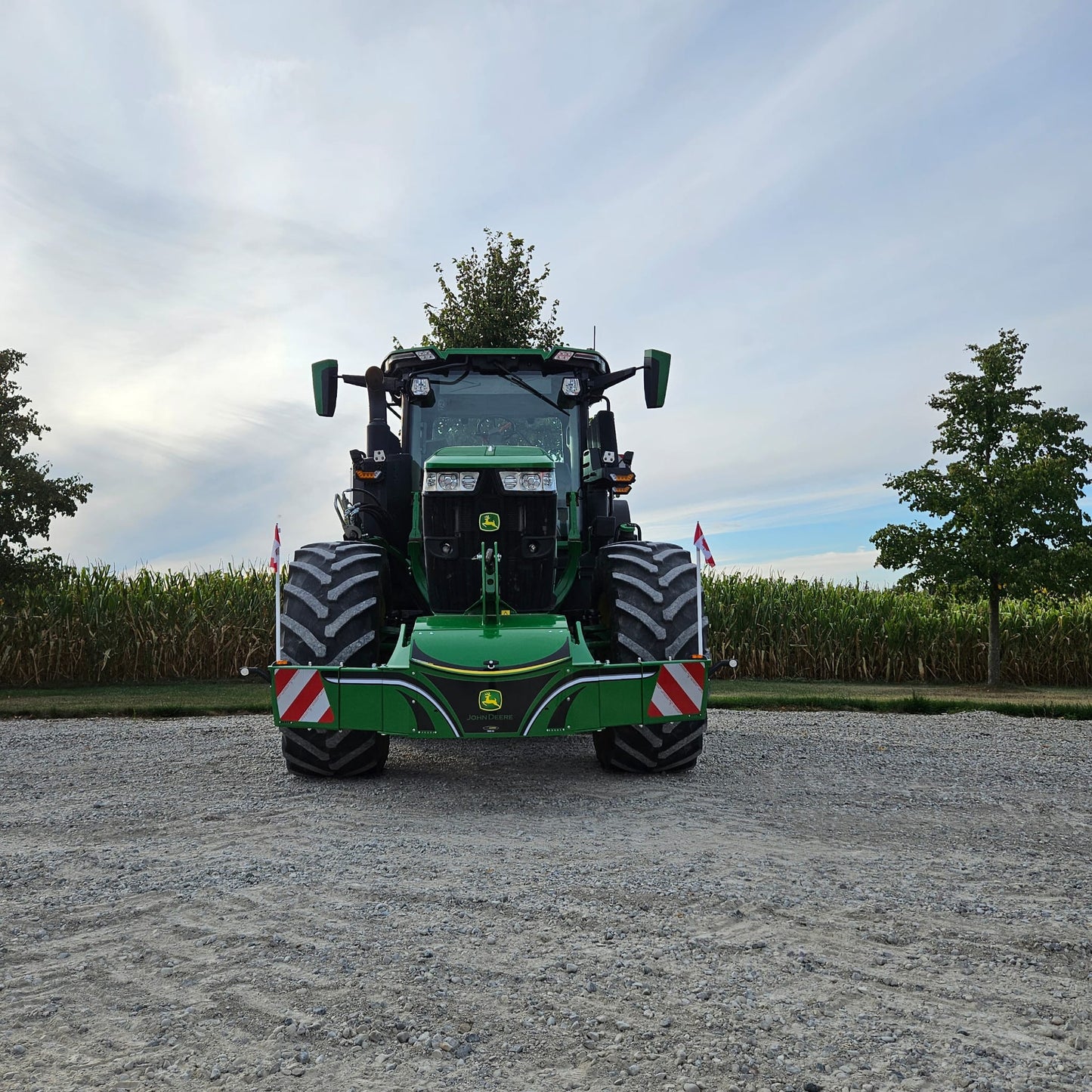 A John Deere TractorBumper 1000KG with LED lights is parked on gravel before a cornfield under cloudy skies, flanked by trees and Danish flags.