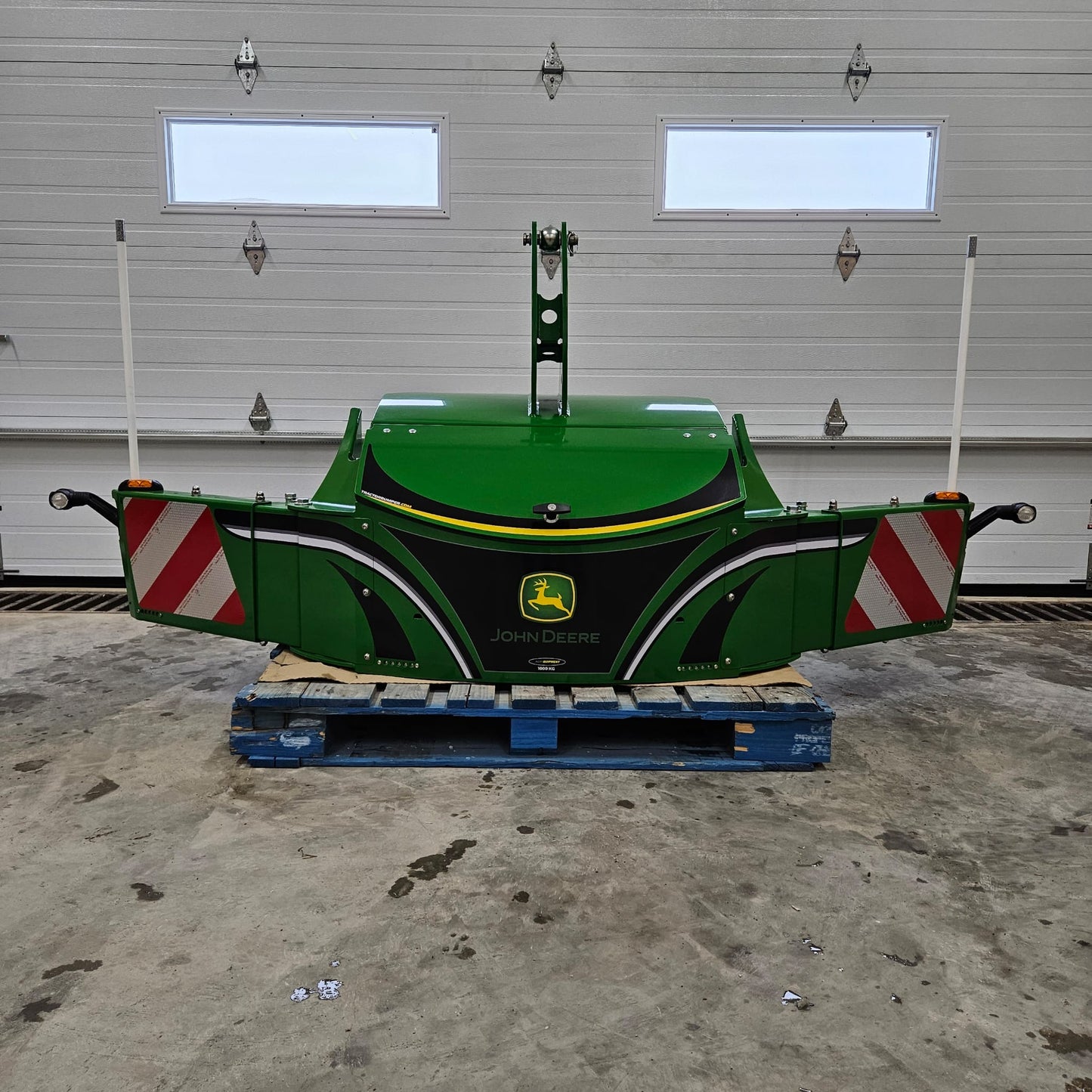 A green John Deere TractorBumper 1000KG with adjustable panels rests on a blue pallet indoors, positioned in front of a closed white garage door featuring two windows.