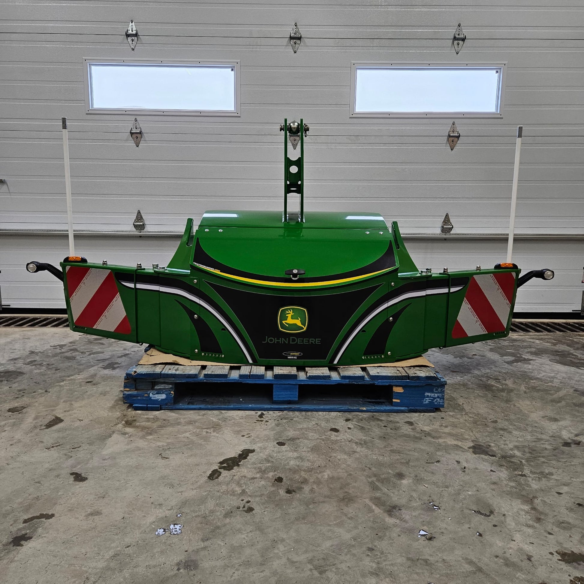 A green John Deere TractorBumper 1000KG with adjustable panels rests on a blue pallet indoors, positioned in front of a closed white garage door featuring two windows.