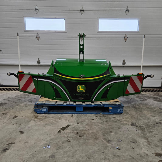 A green John Deere TractorBumper 1000KG with adjustable panels rests on a blue pallet indoors, positioned in front of a closed white garage door featuring two windows.