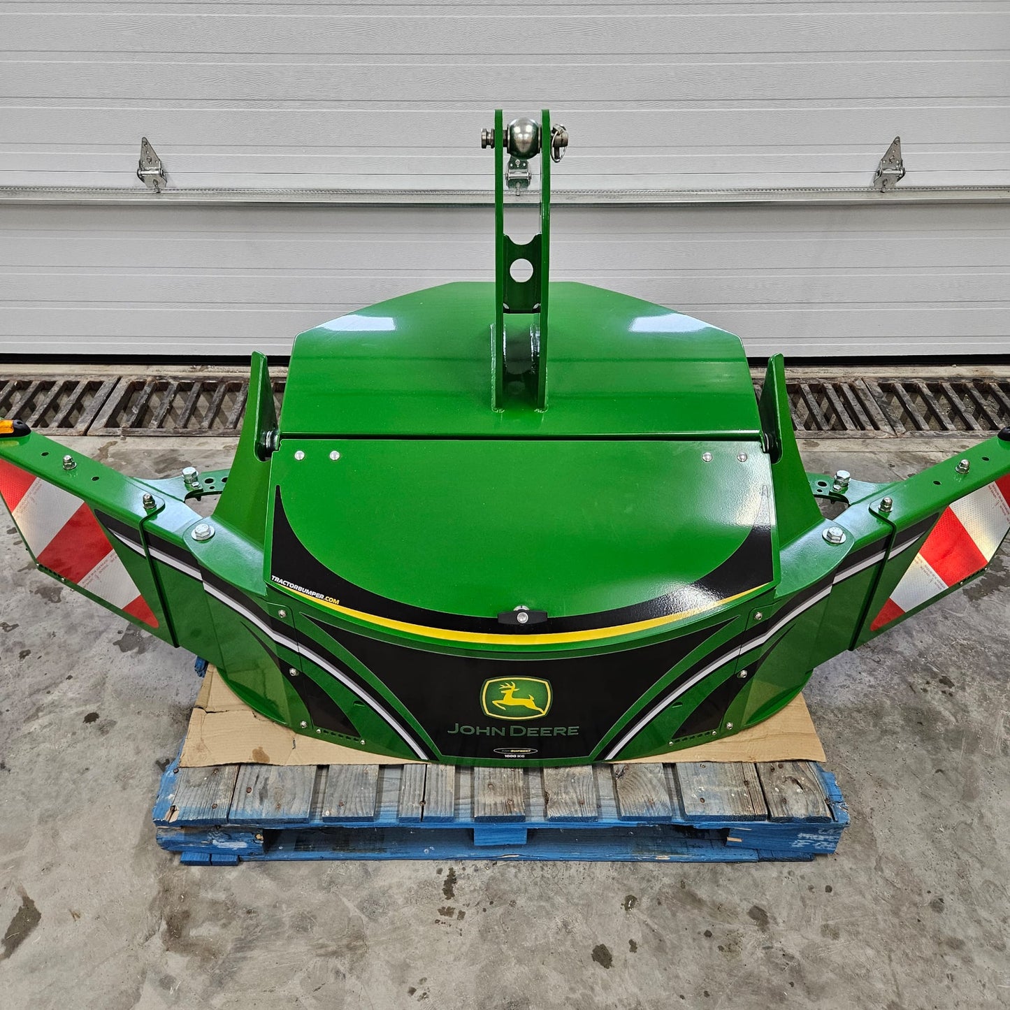 A green John Deere TractorBumper 1000KG with red and white reflective stripes sits on a blue pallet before a white garage door, featuring adjustable panels for added versatility.