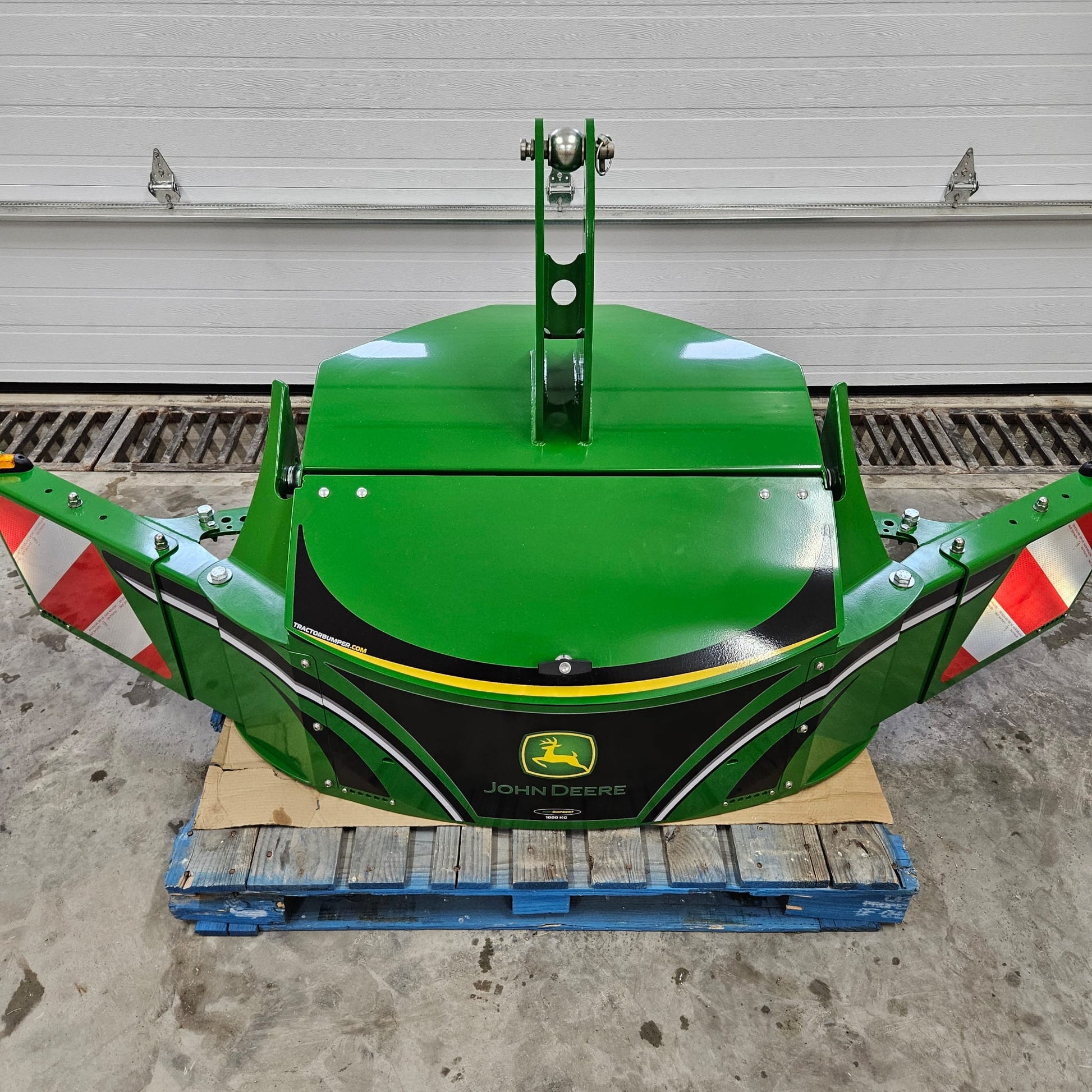 A green John Deere TractorBumper 1000KG with red and white reflective stripes sits on a blue pallet before a white garage door, featuring adjustable panels for added versatility.