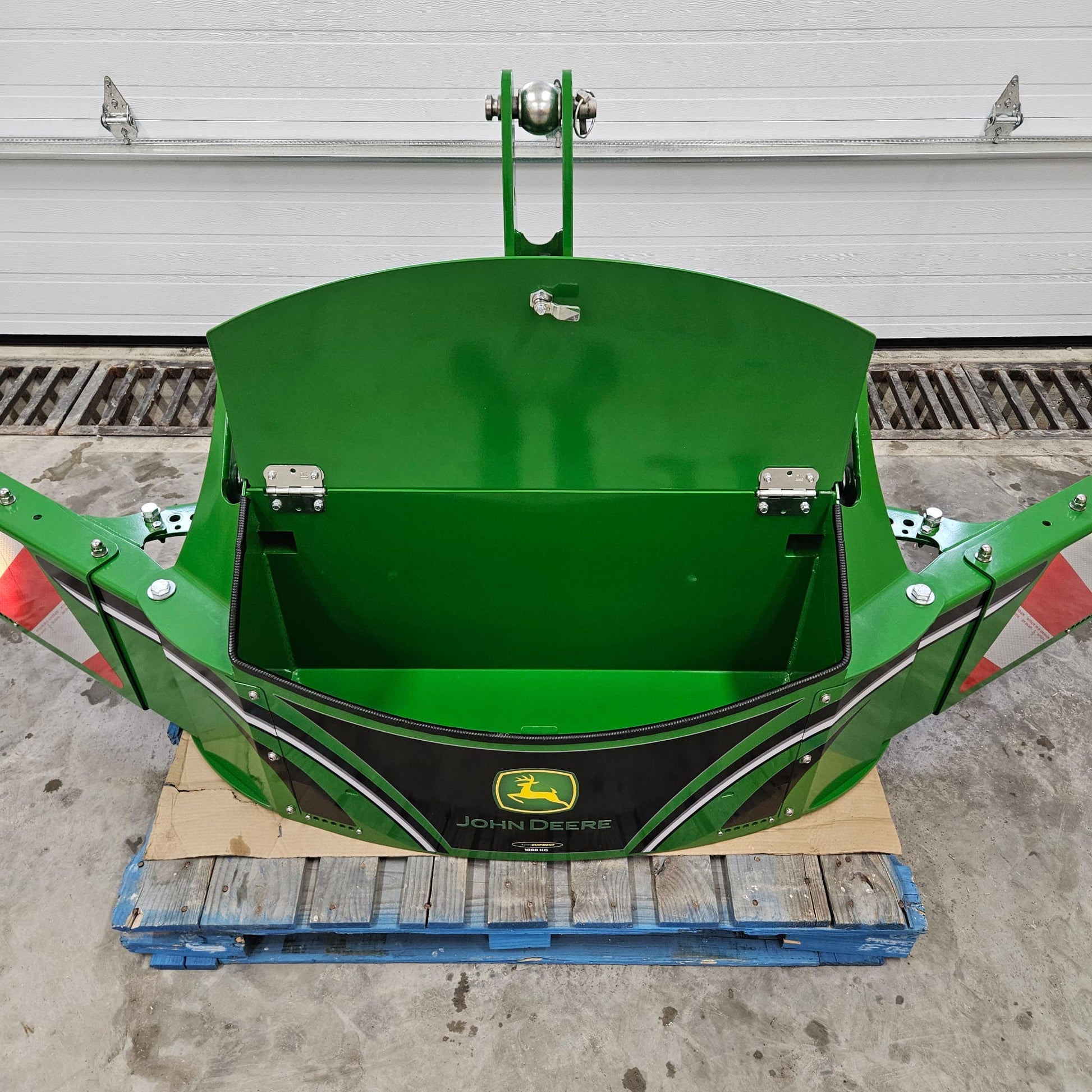 The John Deere TractorBumper 1000KG, a green attachment with adjustable panels and open lid showing an empty compartment, sits on a blue pallet in a garage. Red reflectors are on each side; the white garage door is closed in the background.