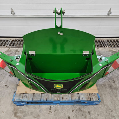 The John Deere TractorBumper 1000KG, a green attachment with adjustable panels and open lid showing an empty compartment, sits on a blue pallet in a garage. Red reflectors are on each side; the white garage door is closed in the background.