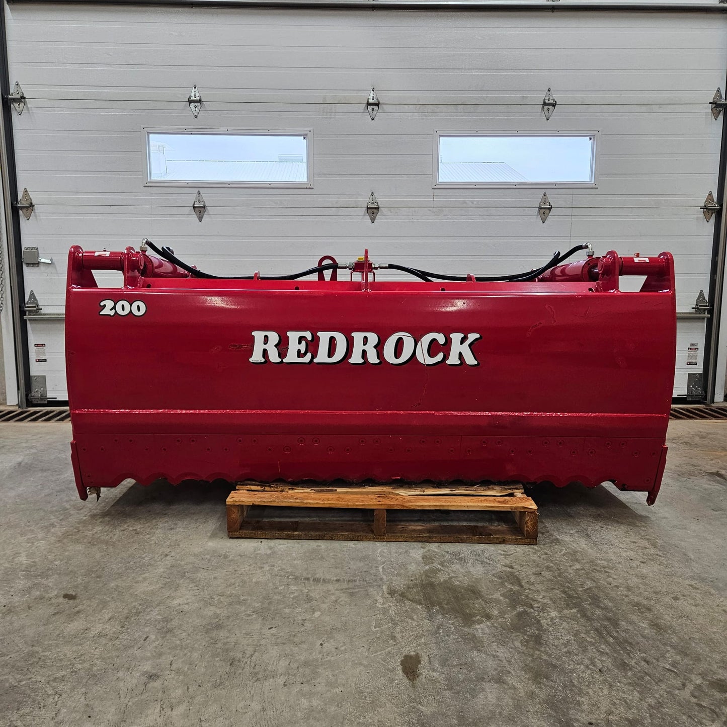An UNUSED REDROCK Shear Bucket 200 (6’6”), a large red heavy-duty silage-cutting implement, sits on a wooden pallet inside a garage with white doors and small windows.