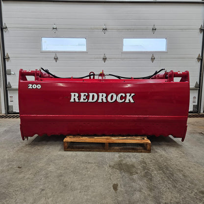 A large red agricultural machine, the Redrock Machinery Shear Bucket Block Cutter, sits on a wooden pallet inside a garage with white doors and two windows.