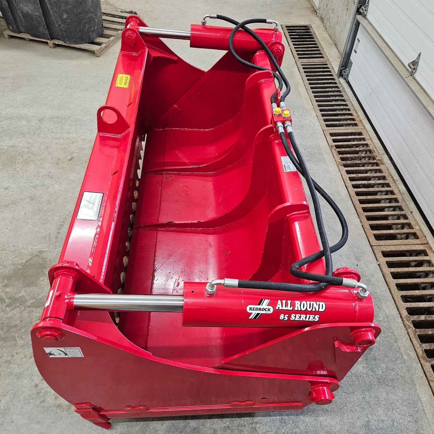 A bright red REDROCK Shear Bucket 200 (6’6”) | UNUSED hydraulic attachment is shown indoors on a concrete floor, featuring visible pistons, hoses, and metal parts—ideal for silage cutting—beside a grated floor drain and white garage door.