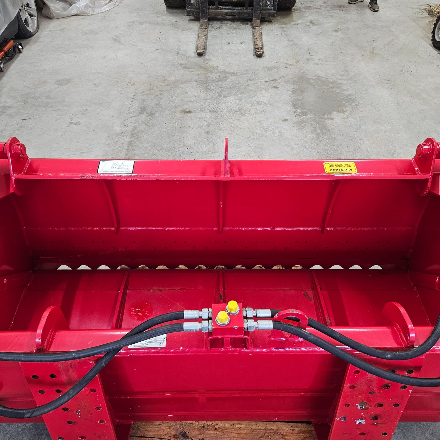 A close-up of the UNUSED REDROCK Shear Bucket 200 (6’6”) shows its bright red, heavy-duty build, hydraulic hoses, and warning labels on a concrete floor, with forklift forks and a person’s legs suggesting silage cutting use.