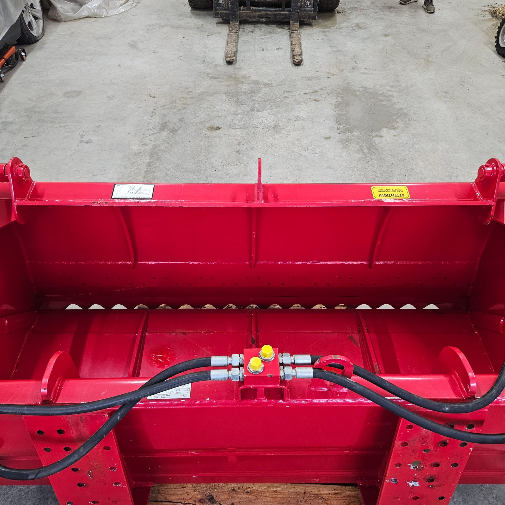 A close-up of the UNUSED REDROCK Shear Bucket 200 (6’6”) shows its bright red, heavy-duty build, hydraulic hoses, and warning labels on a concrete floor, with forklift forks and a person’s legs suggesting silage cutting use.
