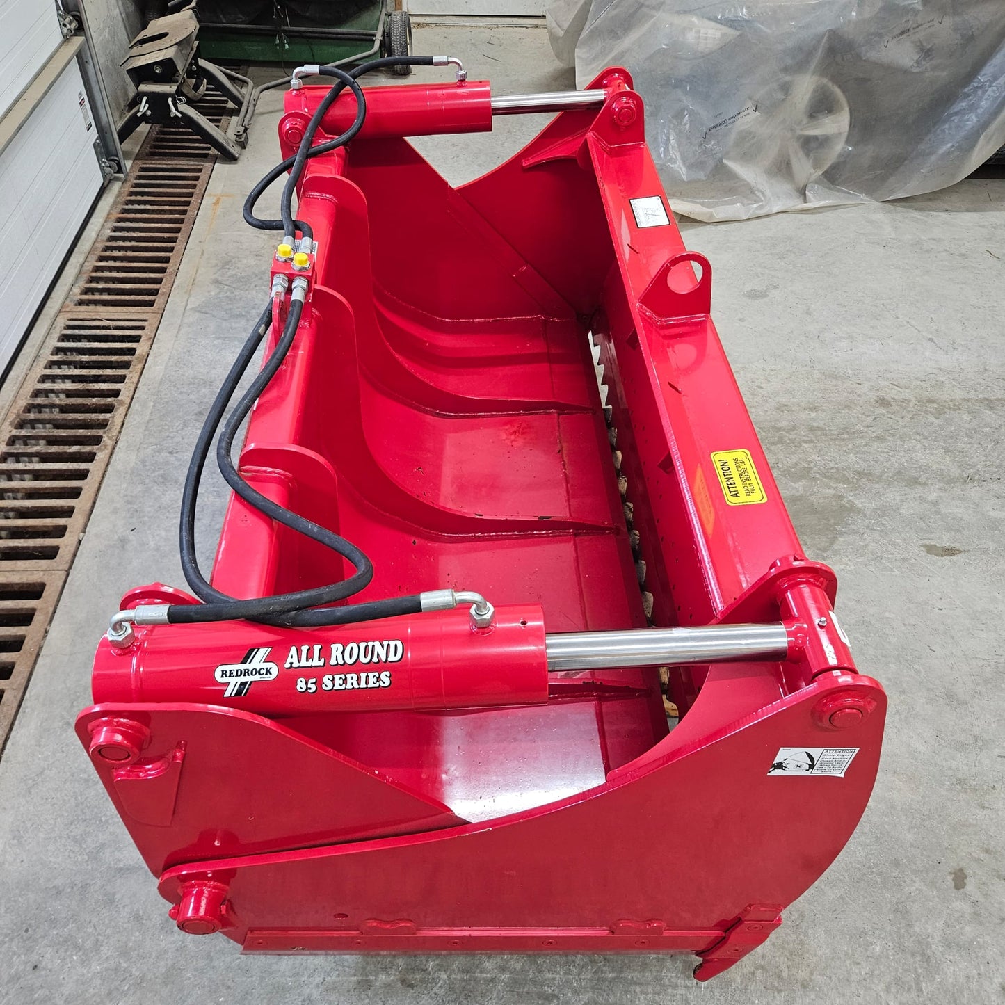 A bright red UNUSED REDROCK Shear Bucket 200 (6’6”) sits on a concrete workshop floor, featuring hydraulic hoses and warning decals.