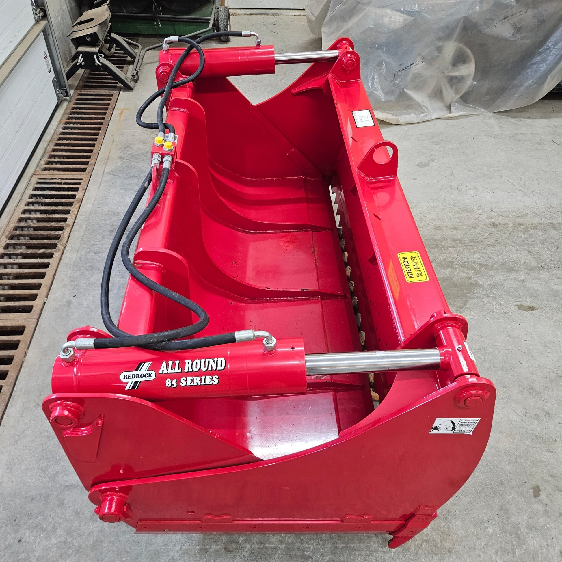 A bright red UNUSED REDROCK Shear Bucket 200 (6’6”) sits on a concrete workshop floor, featuring hydraulic hoses and warning decals.