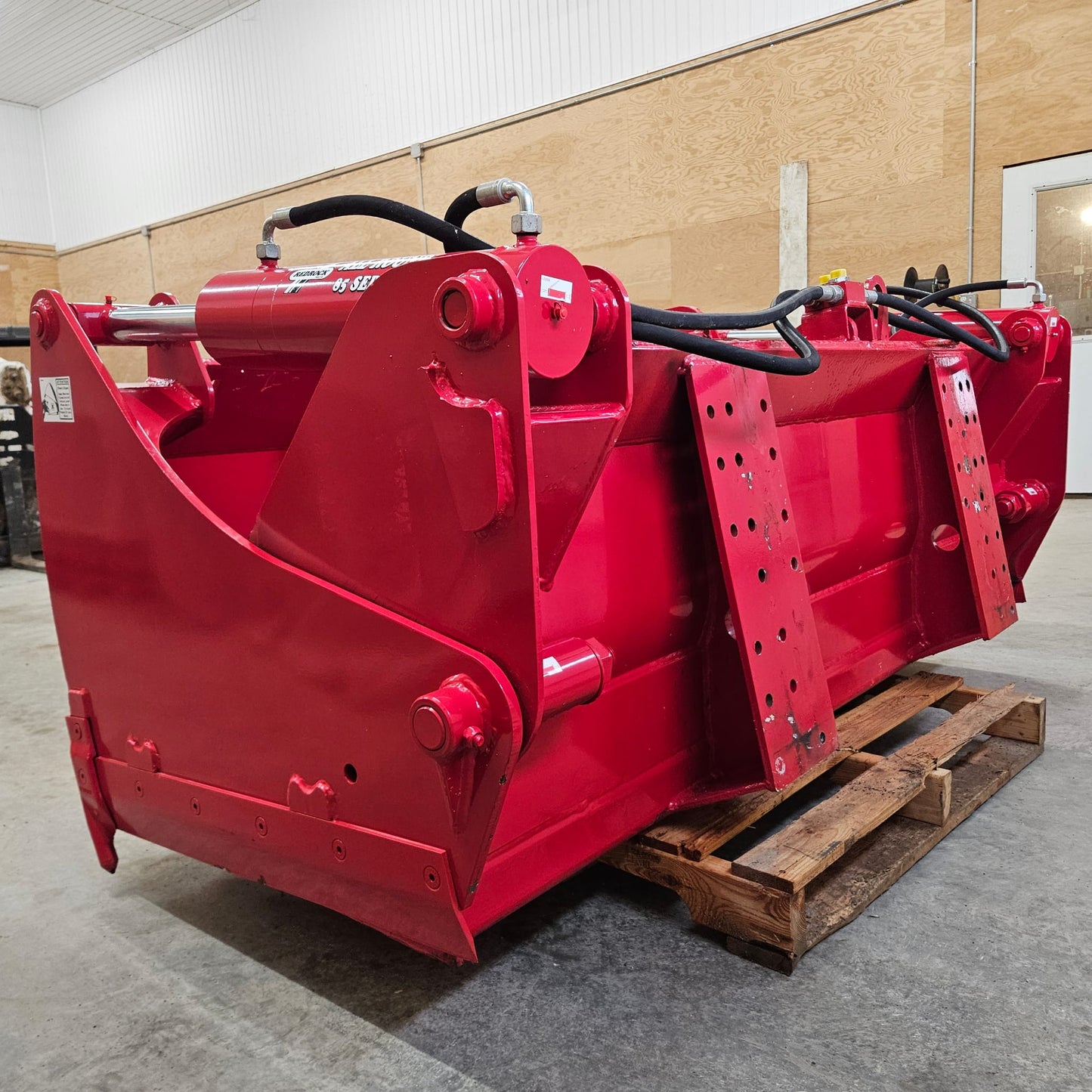 An UNUSED REDROCK Shear Bucket 200 (6’6”), bright red and heavy-duty with hydraulic hoses, sits on a wooden pallet inside a warehouse with light-colored walls and a concrete floor.