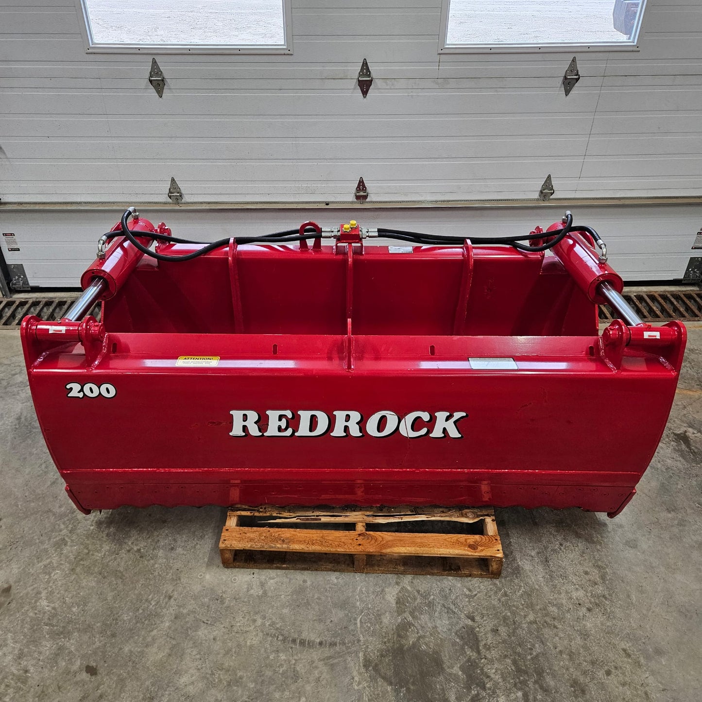 An UNUSED REDROCK Shear Bucket 200 (6’6”) sits on a wooden pallet in front of a white garage door inside a building, its visible hydraulic hoses highlighting its robust design for silage cutting.