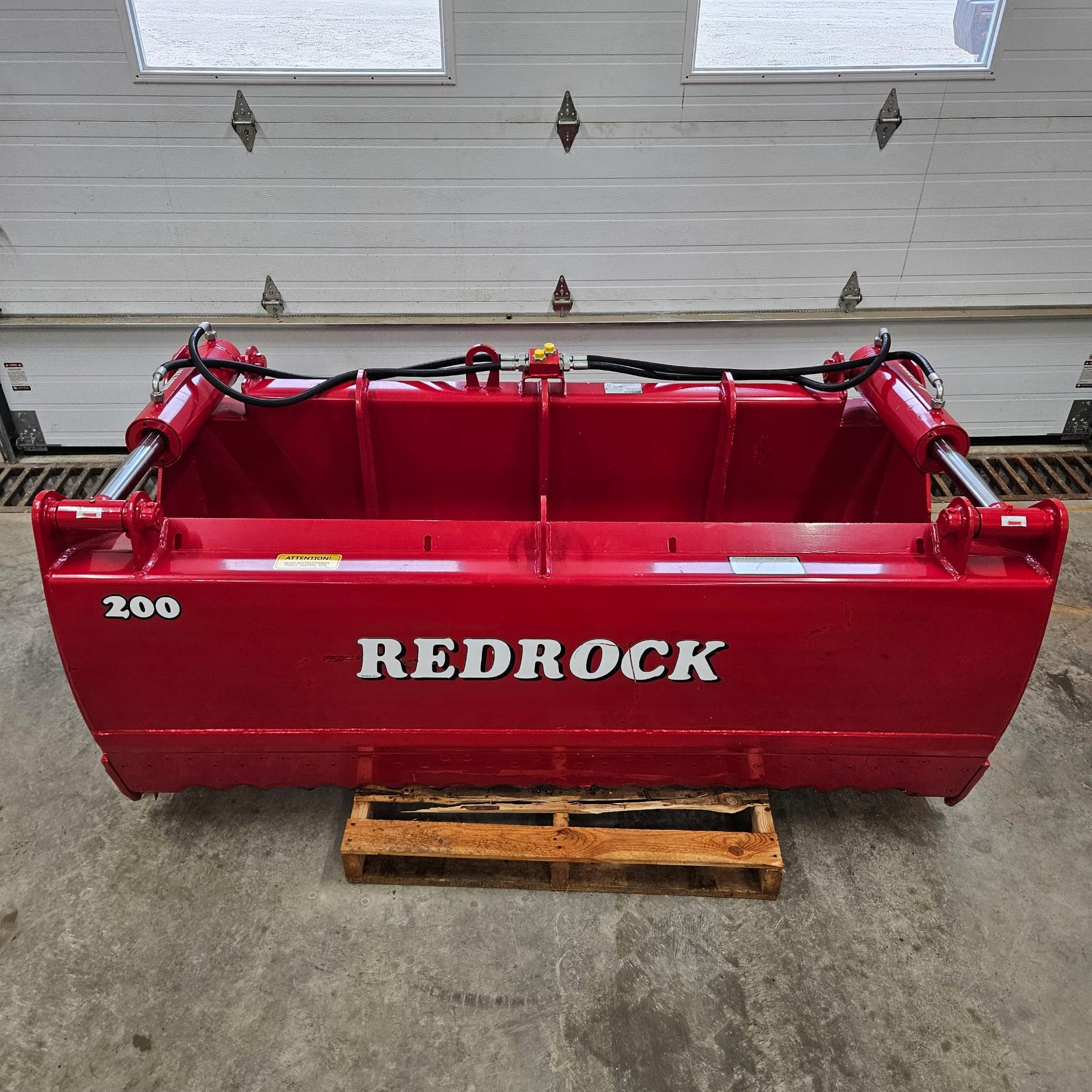 An UNUSED REDROCK Shear Bucket 200 (6’6”) sits on a wooden pallet in front of a white garage door inside a building, its visible hydraulic hoses highlighting its robust design for silage cutting.