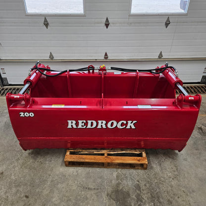 A large red industrial attachment, identified as a Redrock Machinery Shear Bucket Block Cutter, sits on a wooden pallet in a garage with closed white doors in the background.