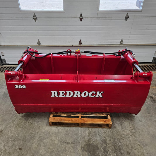 An UNUSED REDROCK Shear Bucket 200 (6’6”) sits on a wooden pallet in front of a white garage door inside a building, its visible hydraulic hoses highlighting its robust design for silage cutting.