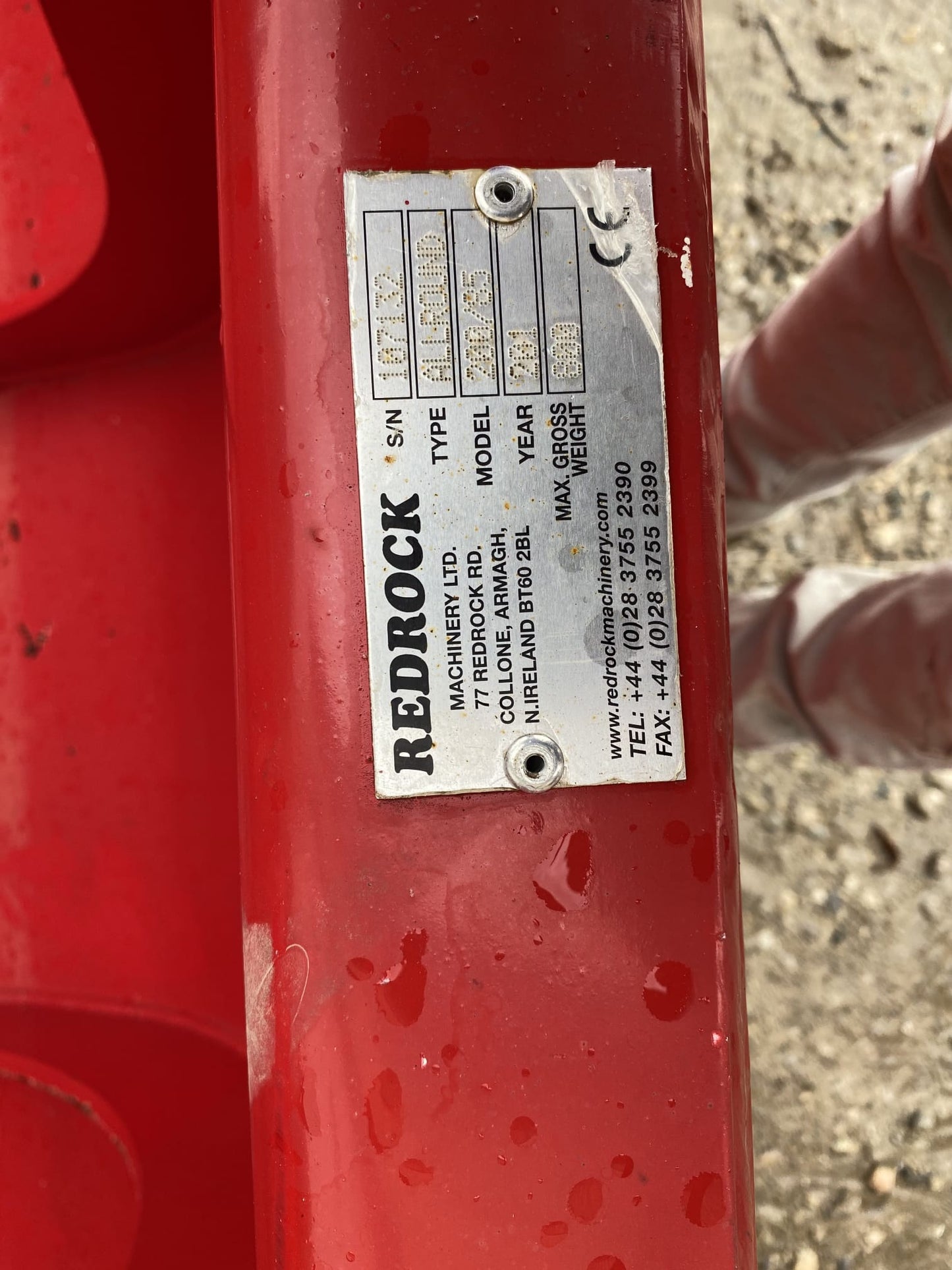 A close-up of the metal ID plate on a REDROCK Shear Bucket 200 (6’6”) UNUSED shows details for REDROCK Machinery Ltd. A person’s legs appear in the background—equipment ideal for silage cutting.